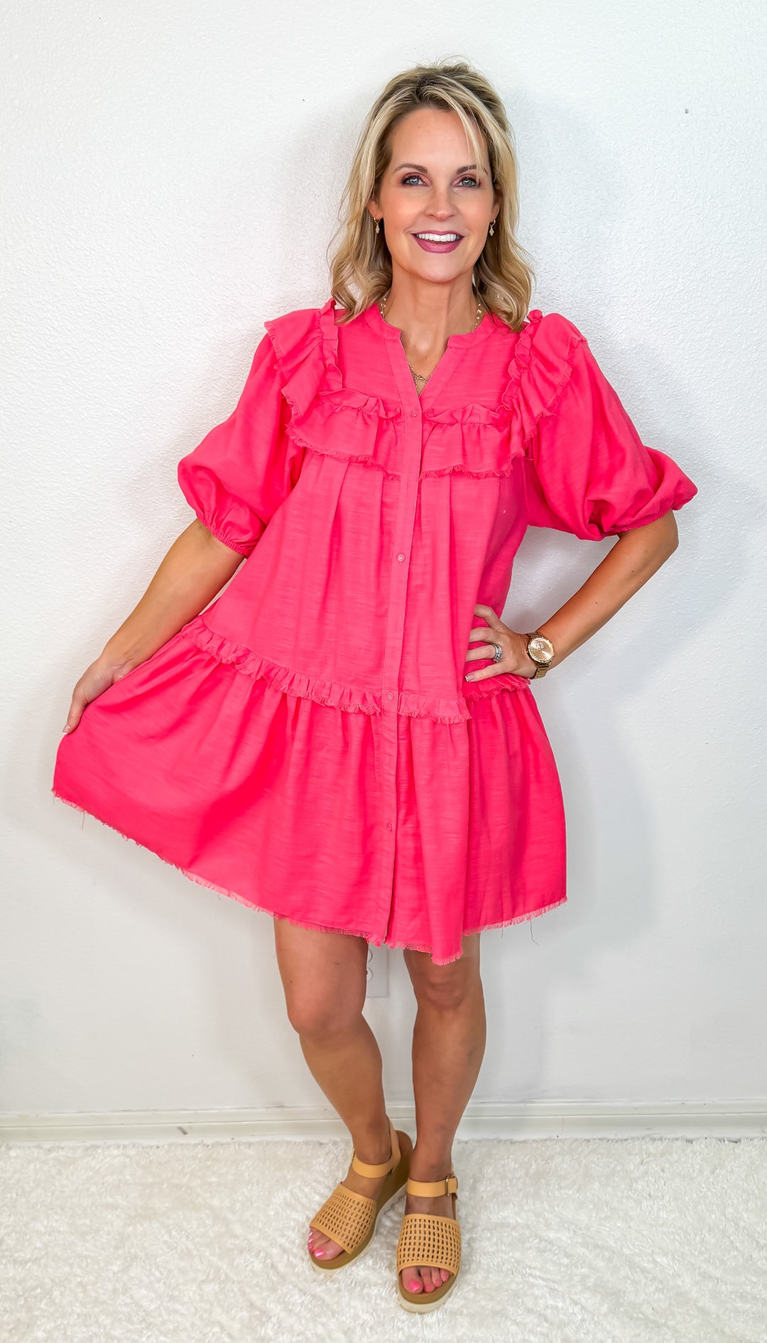 A model wearing a short-sleeved pink dress with a button-up front and ruffle detailing, paired with sandals.