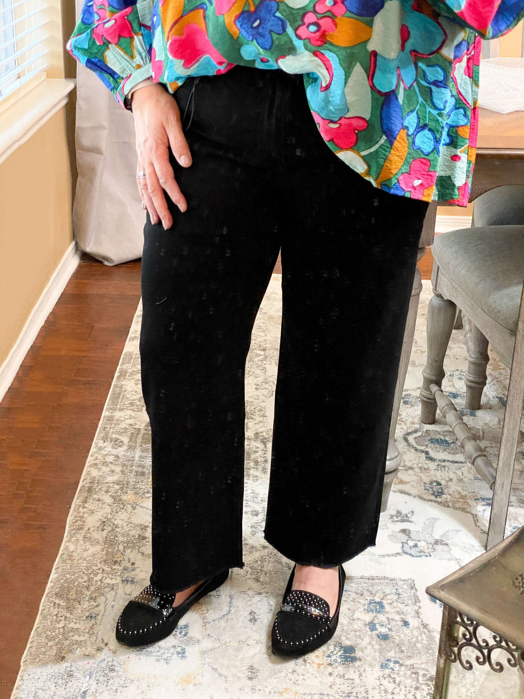 A person wearing black wide-leg crop jeans and a colorful top, standing indoors.