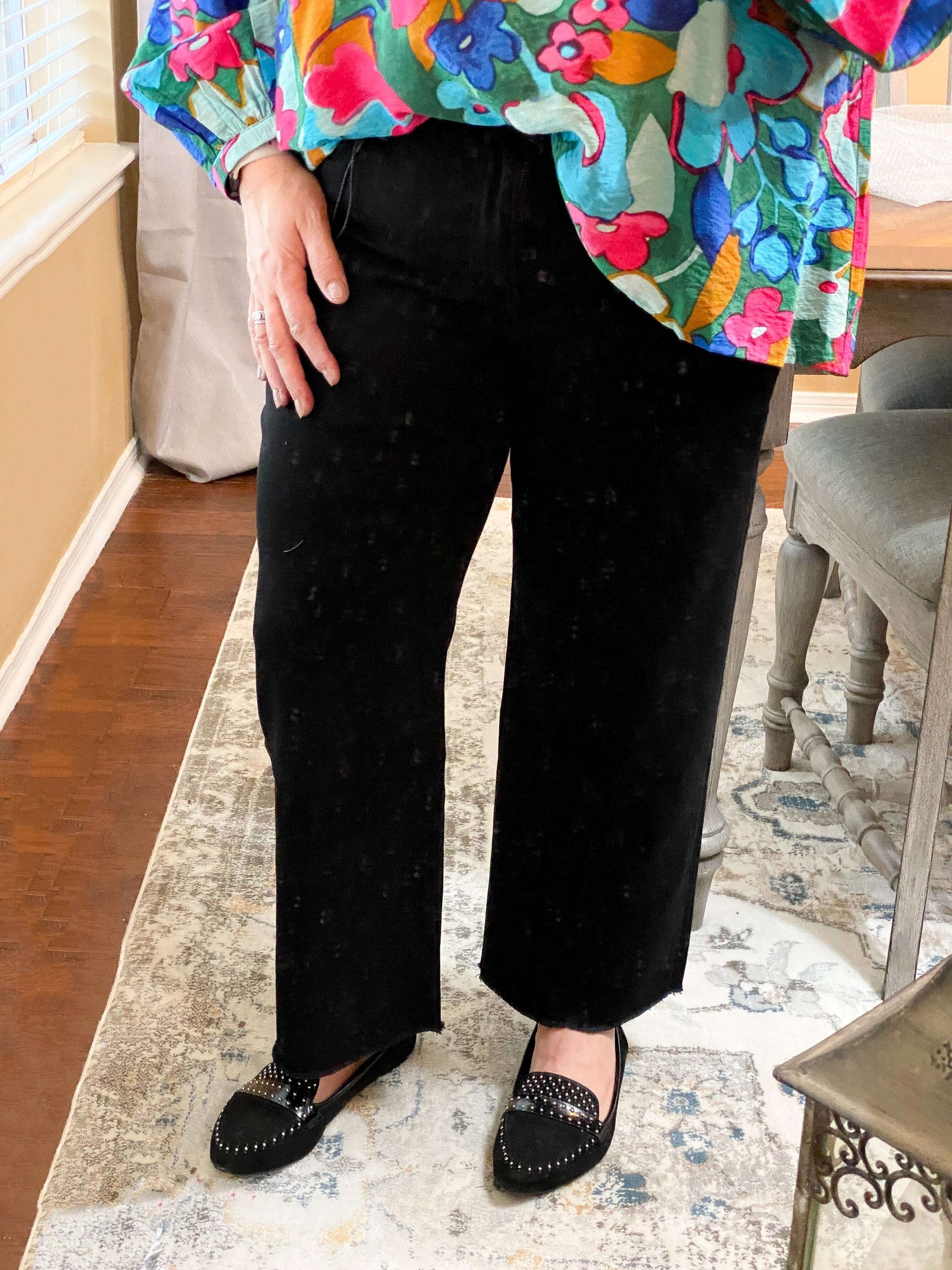 A person wearing black wide-leg crop jeans and a colorful top, standing indoors.