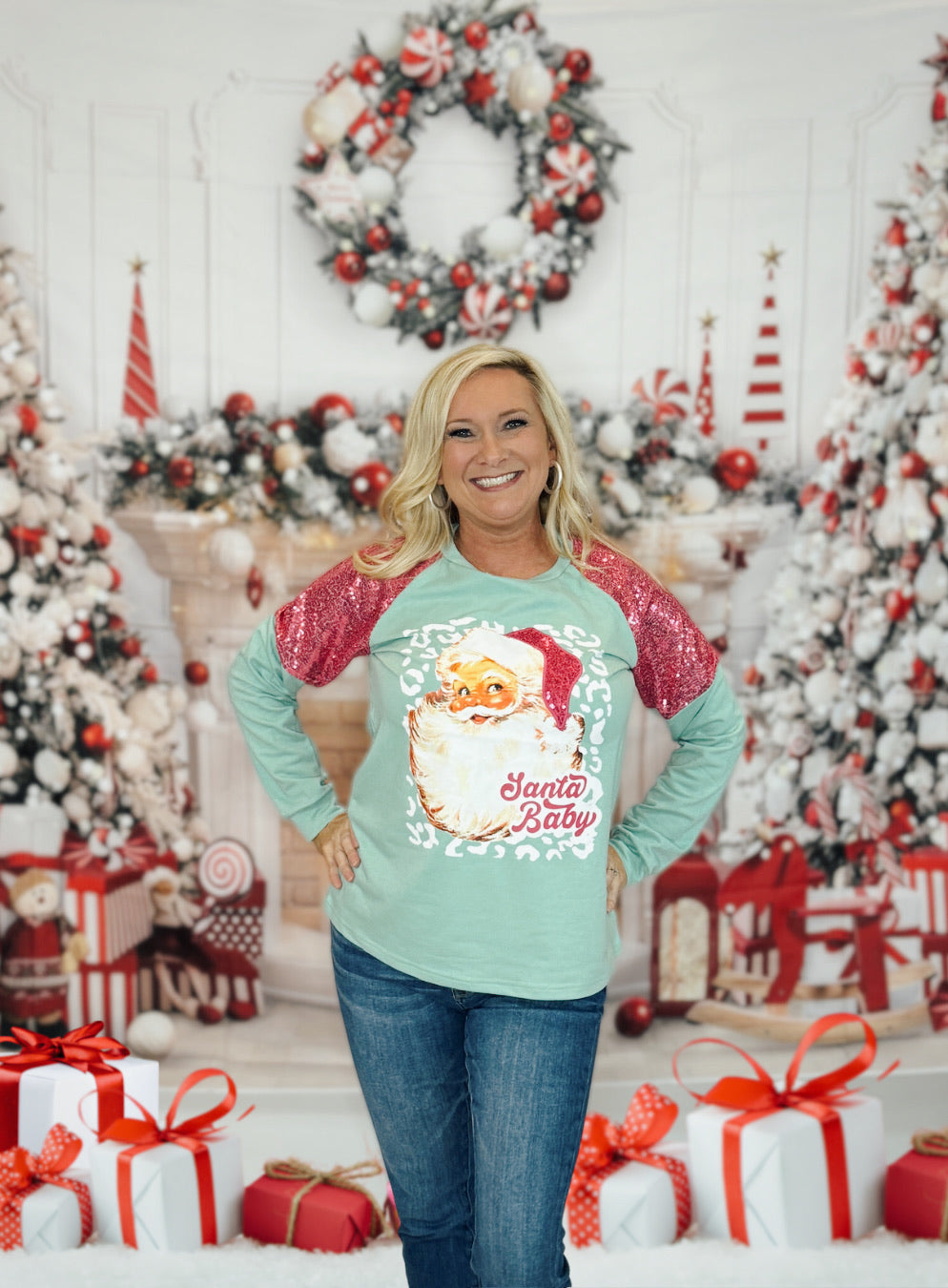 A woman standing in front of a Christmas-themed backdrop wearing a light blue Santa Baby glitzy sweatshirt with a Santa graphic and the text 'Santa Baby' on it.