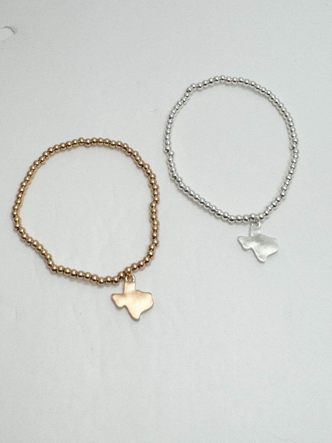 Two bracelets, one gold and one silver, with Texas-shaped charms on a light gray background.