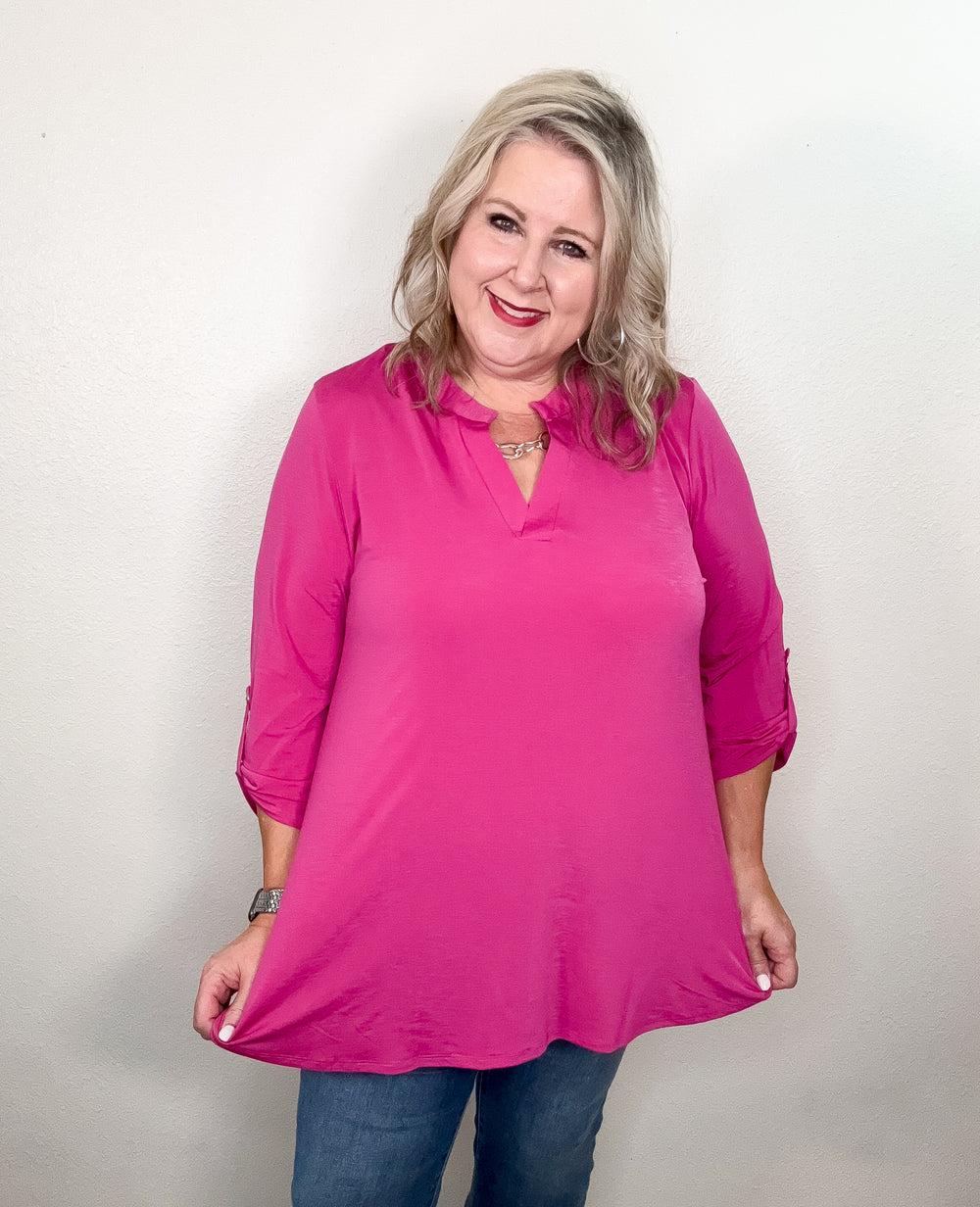 A person wearing a pink top with a notch neckline and 3/4 length sleeves with tab detail, paired with jeans.