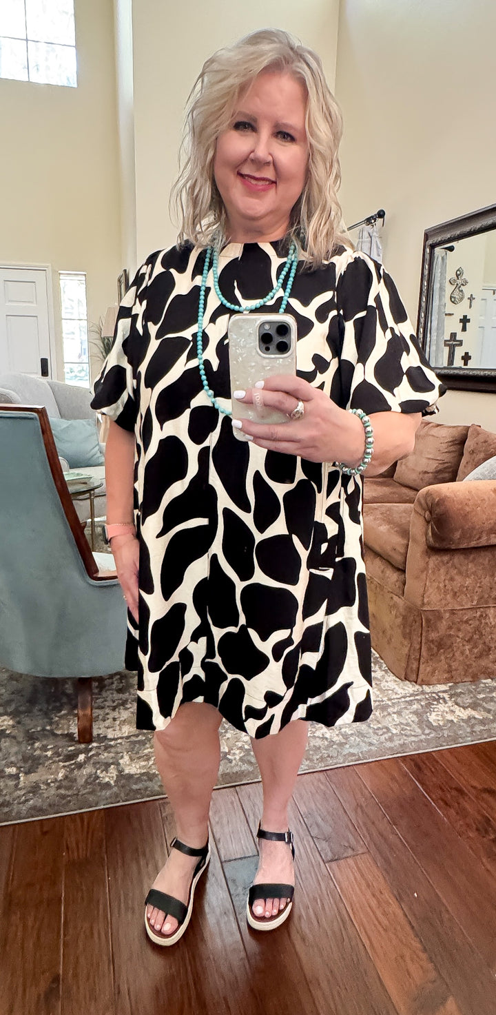 A woman standing in a room wearing a high contrast dress with a bold black and ivory abstract print, featuring elegant puff sleeves.