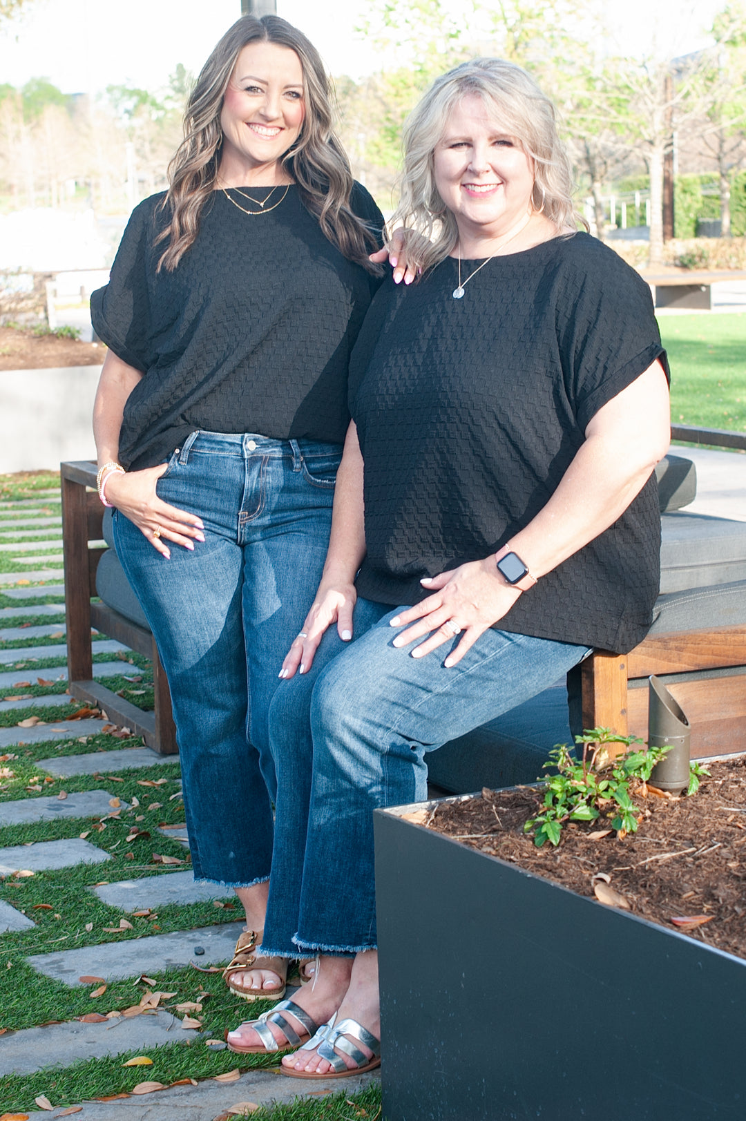 Two women standing next to each other, wearing black boxy tops with a button-down back detail. One woman is wearing glasses, and both are smiling.
