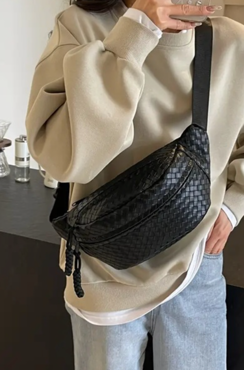 A person wearing a brown sweatshirt and blue jeans is holding a beige crossbody bag with a woven design. The bag is secured to the person's waist.