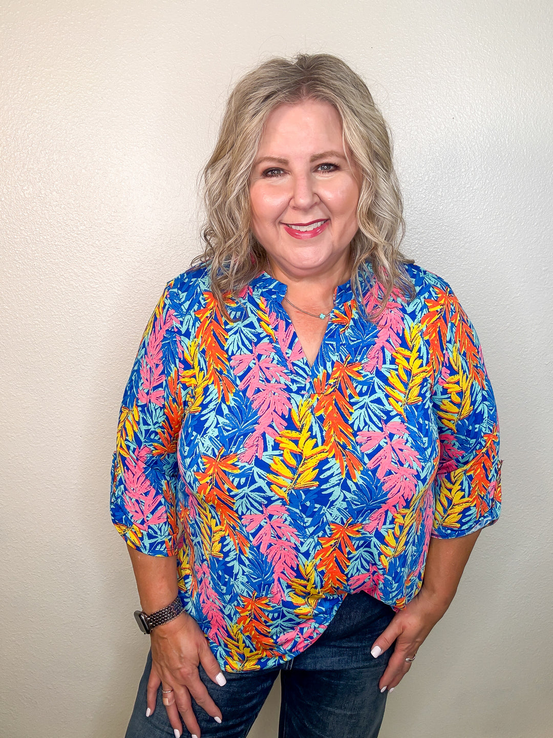A woman smiling and wearing a colorful leafy patterned top with a V-neck and 3/4 length sleeves.