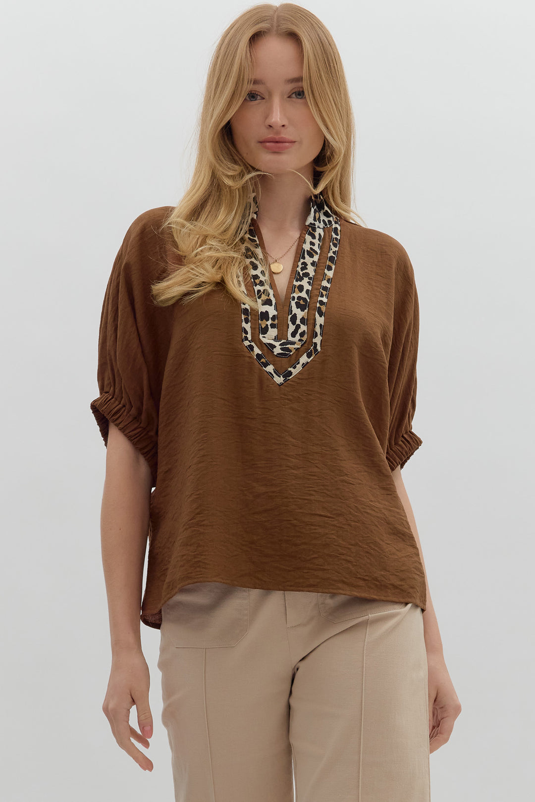 Woman wearing a brown top and beige pants with a leopard print necklace on a white background