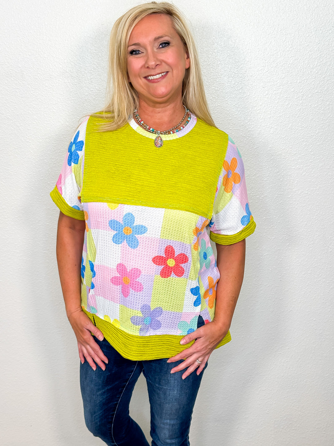 A woman wearing a vibrant top with a chartreuse base and colorful floral details. The top has a relaxed fit with short sleeves, a round neckline, and an hi-low hemline.