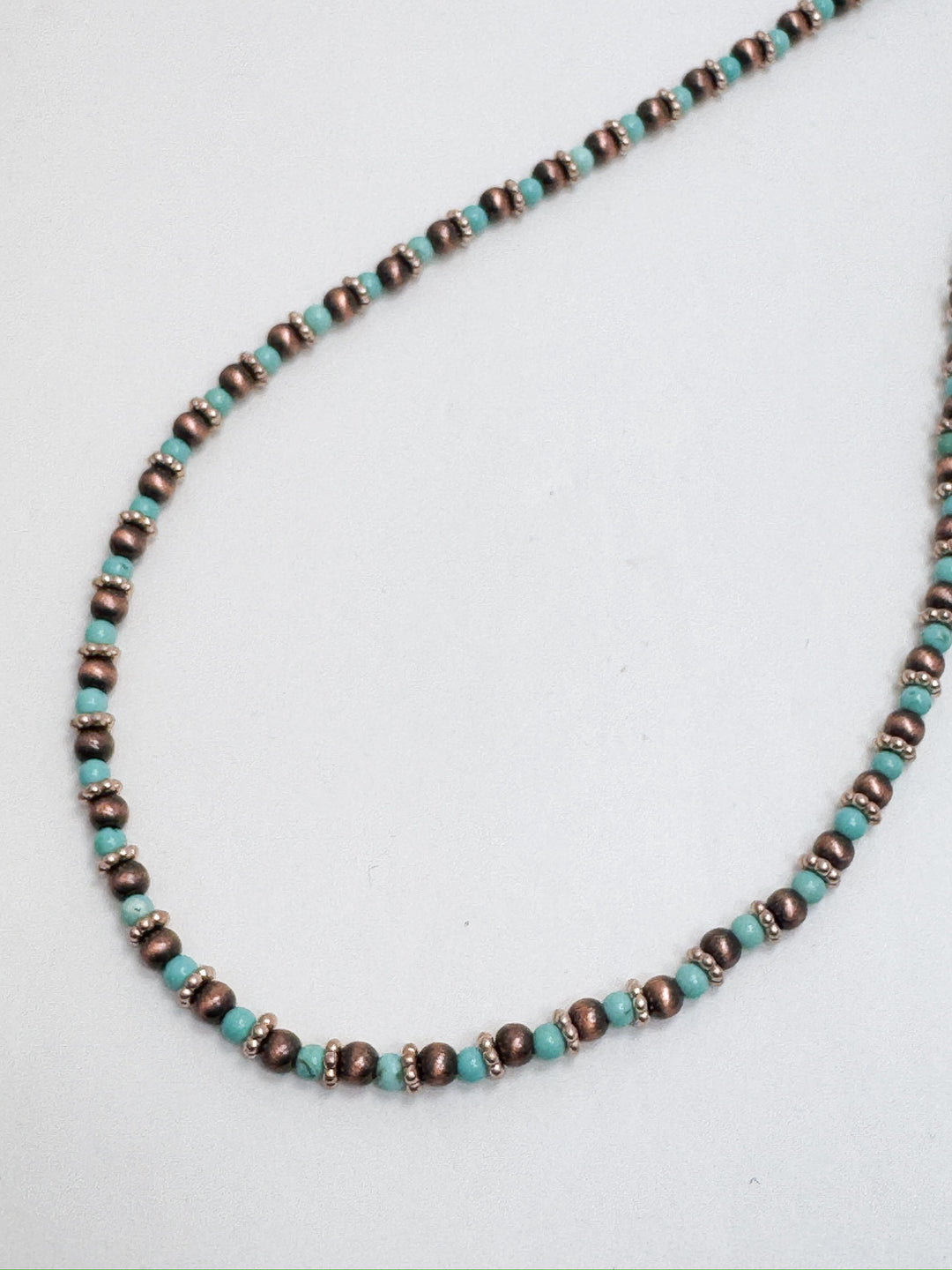 A necklace with a single strand of faux Navajo pearls, featuring turquoise and copper accents, laid out on a plain surface.