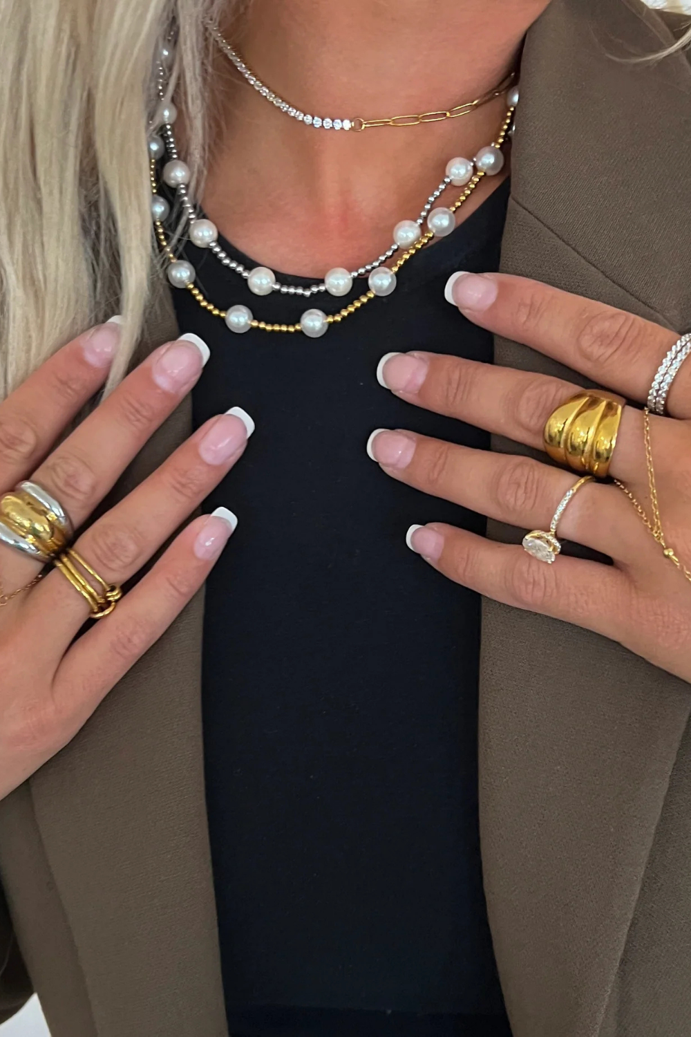 Close-up of a person wearing multiple pearl necklaces, gold rings, and a beige blazer.