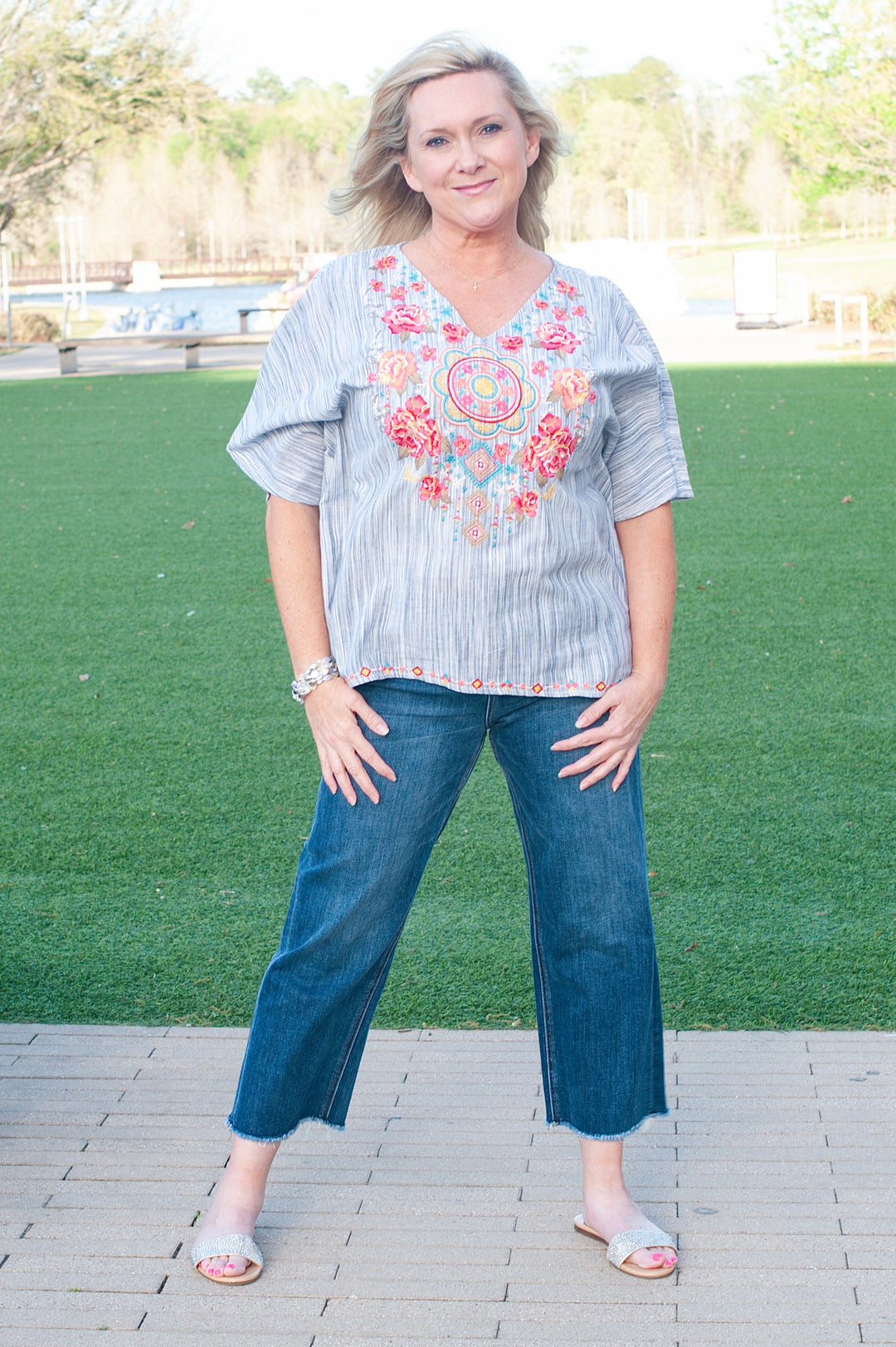 A woman wearing a gray and white striped poncho-style top with floral embroidery, paired with blue jeans and white sandals.