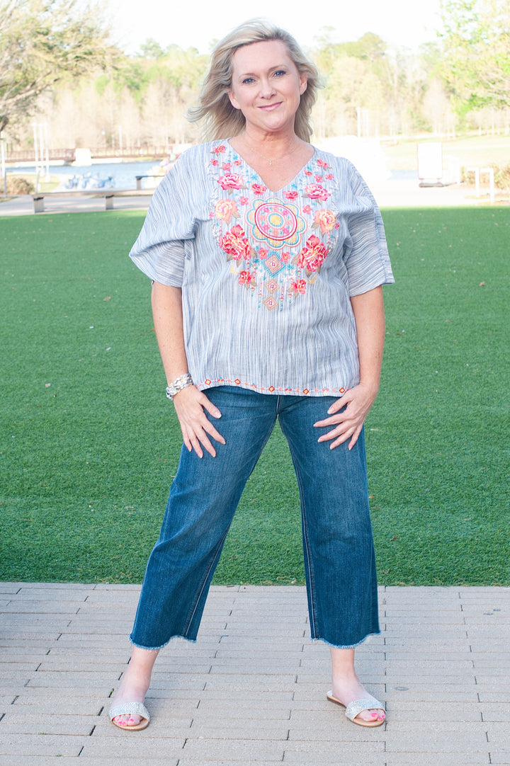A woman wearing a gray and white striped poncho-style top with floral embroidery, paired with blue jeans and white sandals.