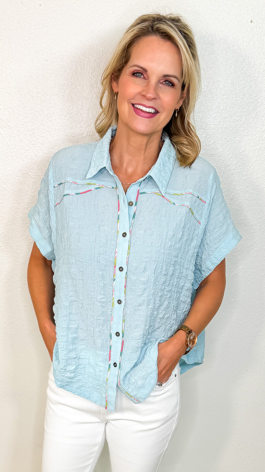 A woman smiling and wearing a blue crinkle-textured top with colorful piping details, short sleeves, and a collared neckline.