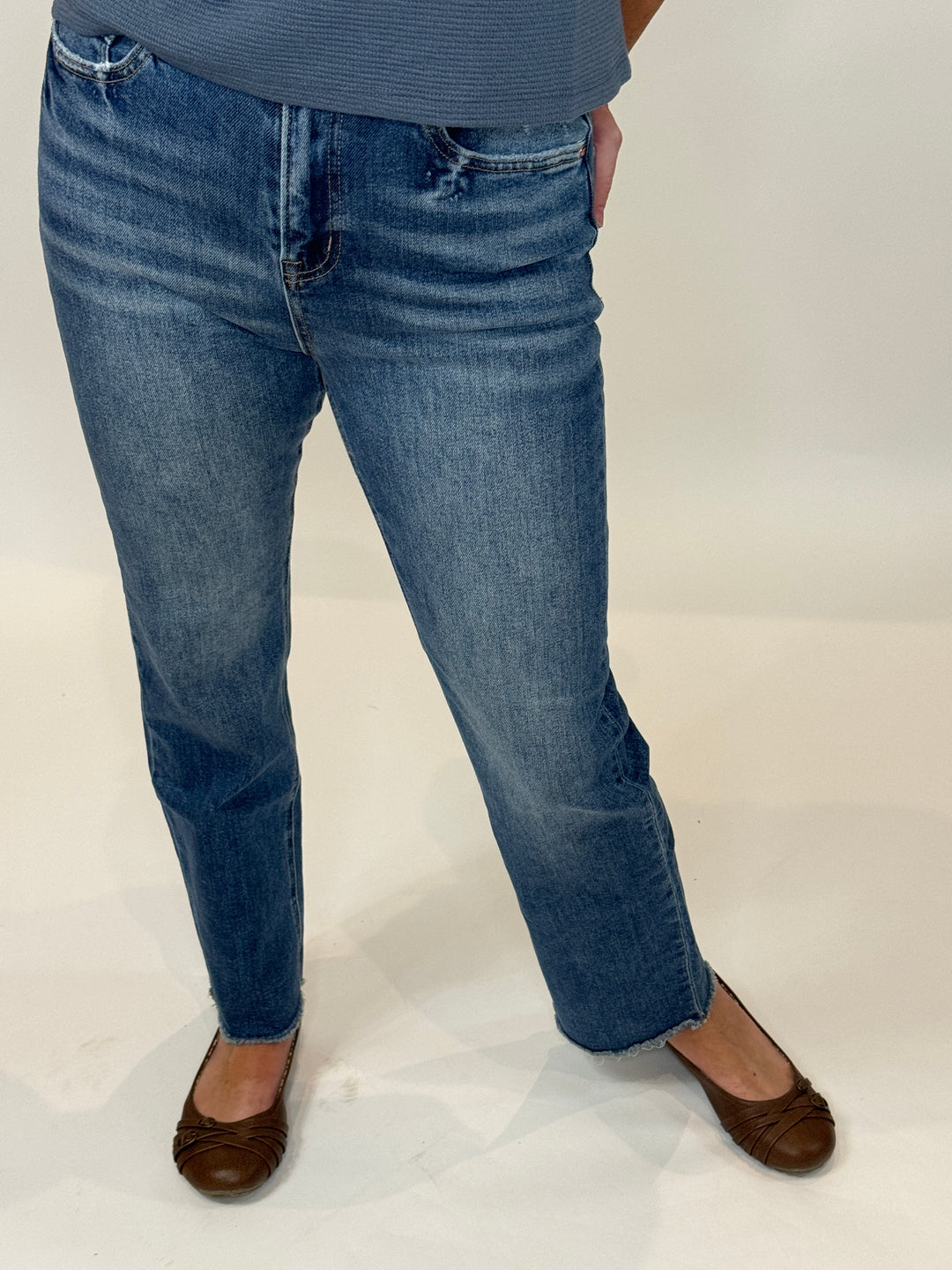 A person standing against a white background, wearing The Alex Jean which is a high-rise straight-leg blue denim jean and brown shoes.
