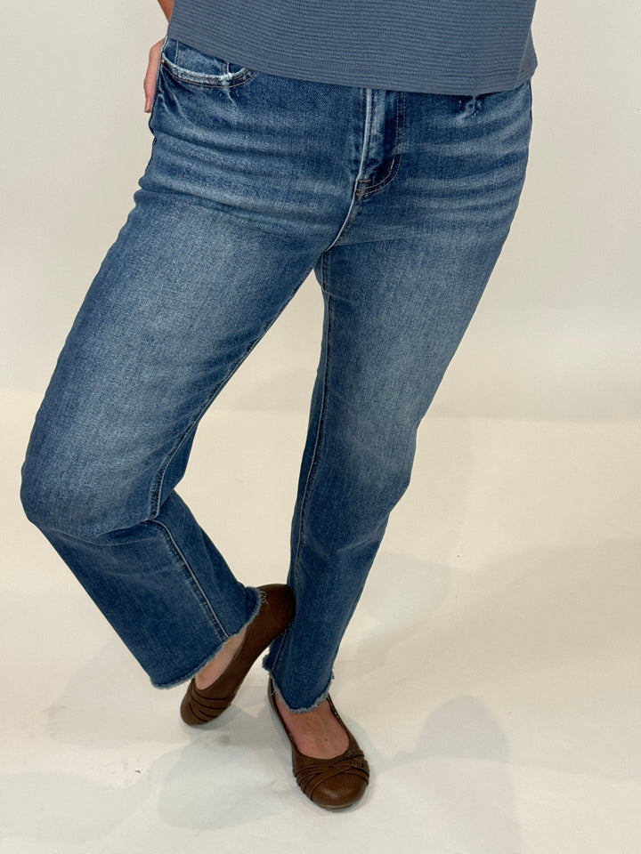 A person standing against a white background, wearing The Alex Jean which is a high-rise straight-leg blue denim jean and brown shoes.