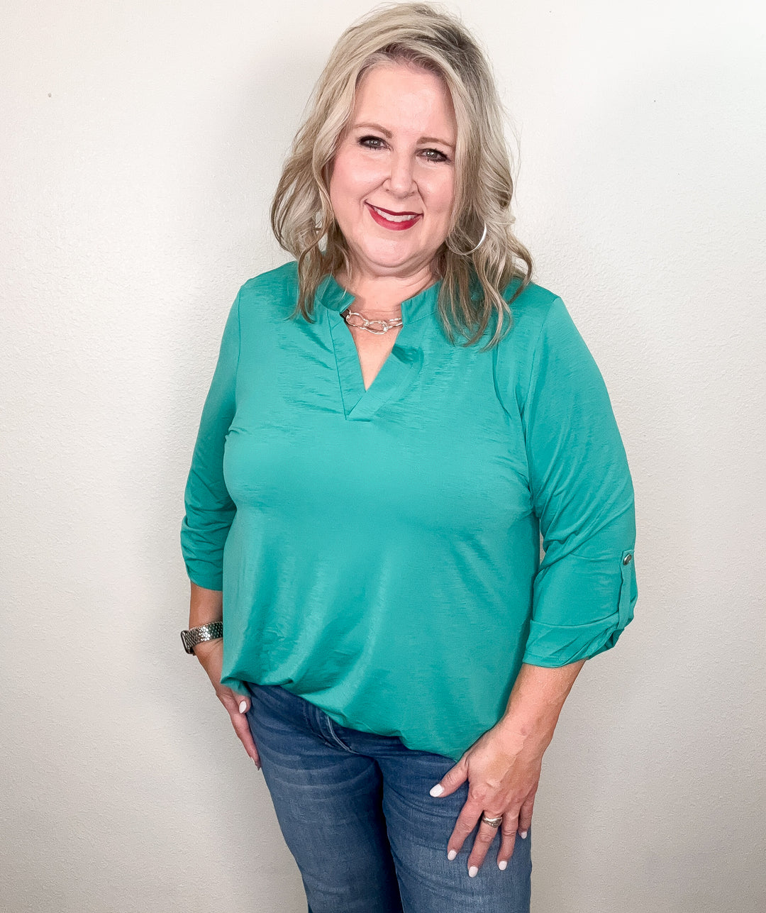 A woman smiling and wearing a green, v-neck, 3/4 sleeve top with a hi-low hemline paired with jeans.