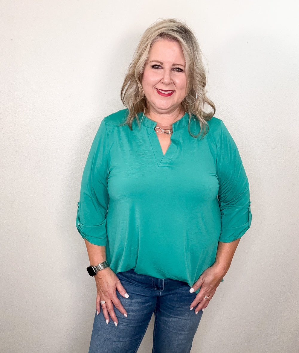 A woman smiling and wearing a green, v-neck, 3/4 sleeve top with a hi-low hemline paired with jeans.