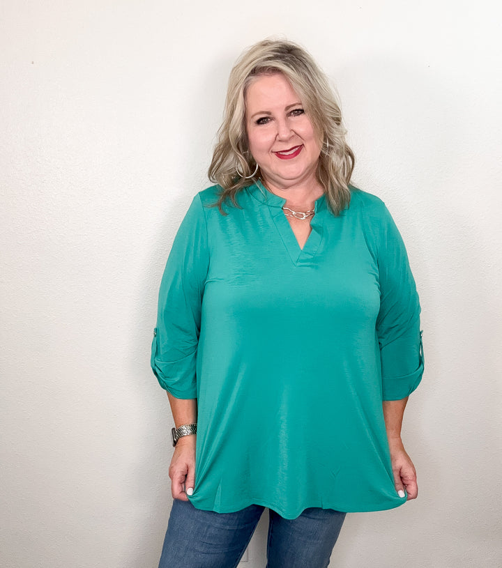 A woman smiling and wearing a green, v-neck, 3/4 sleeve top with a hi-low hemline paired with jeans.