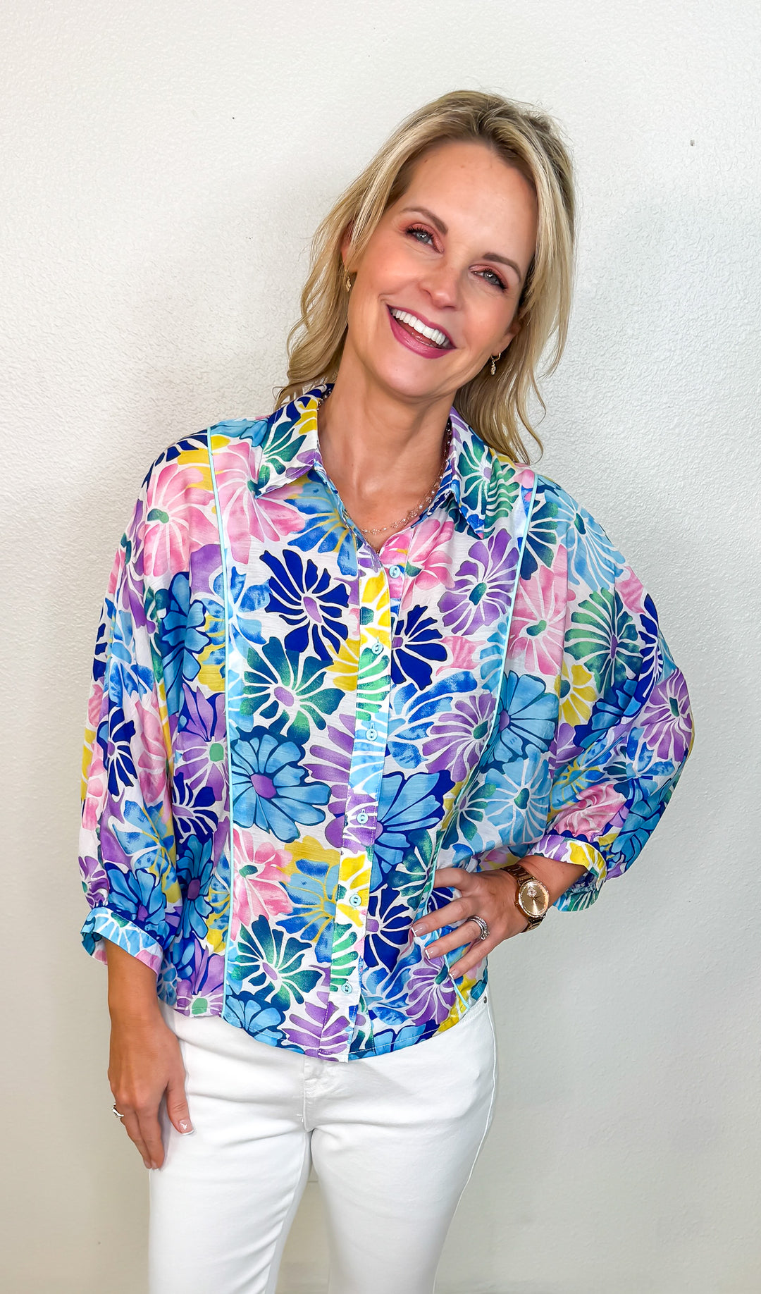 A woman smiling and wearing a vibrant floral print top with 3/4 length sleeves and a collared neckline.