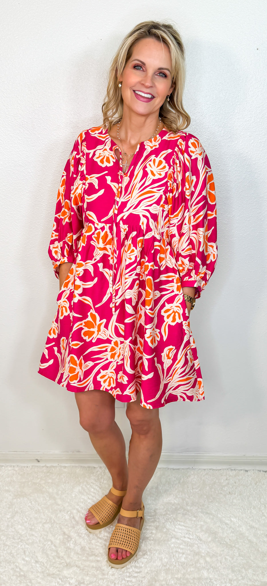 A model wearing a vibrant fuchsia, orange, and cream colored floral dress with a tie keyhole neckline and bubble sleeves.