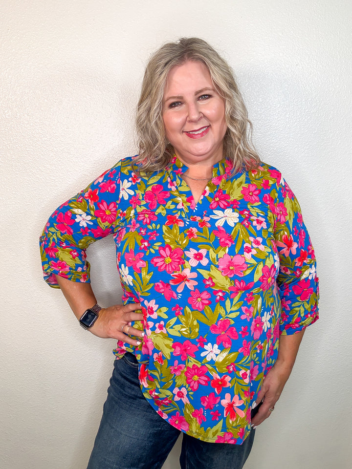 A woman wearing a colorful floral print top with a notch neckline and 3/4 length sleeves with tab detail.