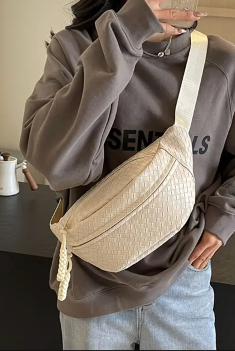 A person wearing a brown sweatshirt and blue jeans is holding a beige crossbody bag with a woven design. The bag is secured to the person's waist.