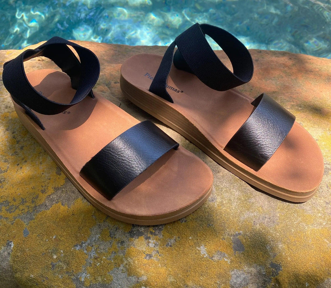 A pair of black and brown sandals with elastic ankle straps, placed on a stone surface near a pool.