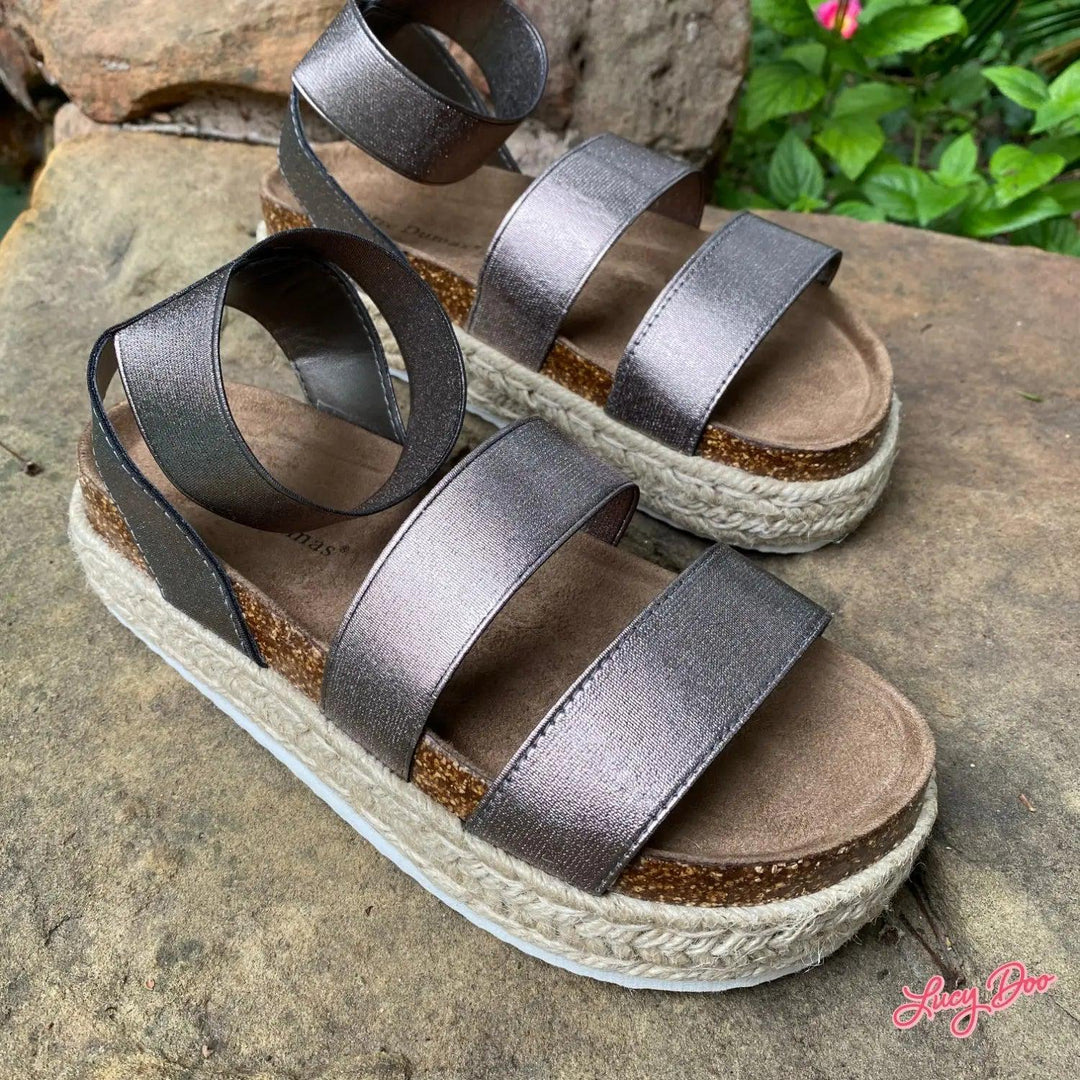 A pair of pewter metallic sandals with a platform sole and three straps, displayed on a rock.