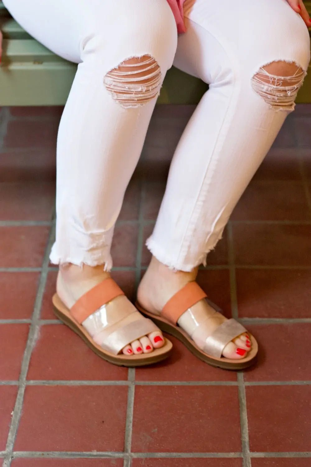 A pair of metallic strapped sandals in coral and clear colors, with a person seated in white pants with ripped details, wearing the sandals.